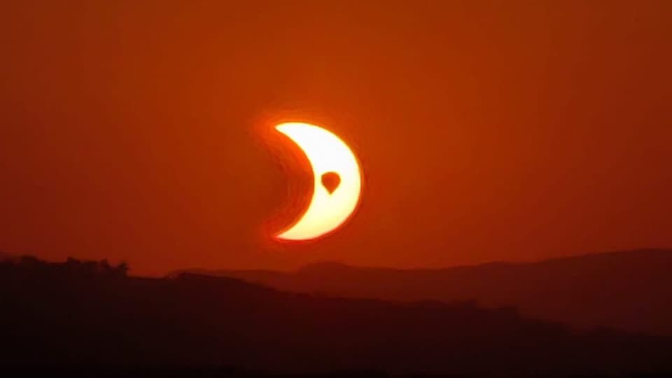 Partial solar eclipse at sunset — a crescent sun glowing deep red-orange low over a dark ridgeline, a small silhouette crossing the sun's face.